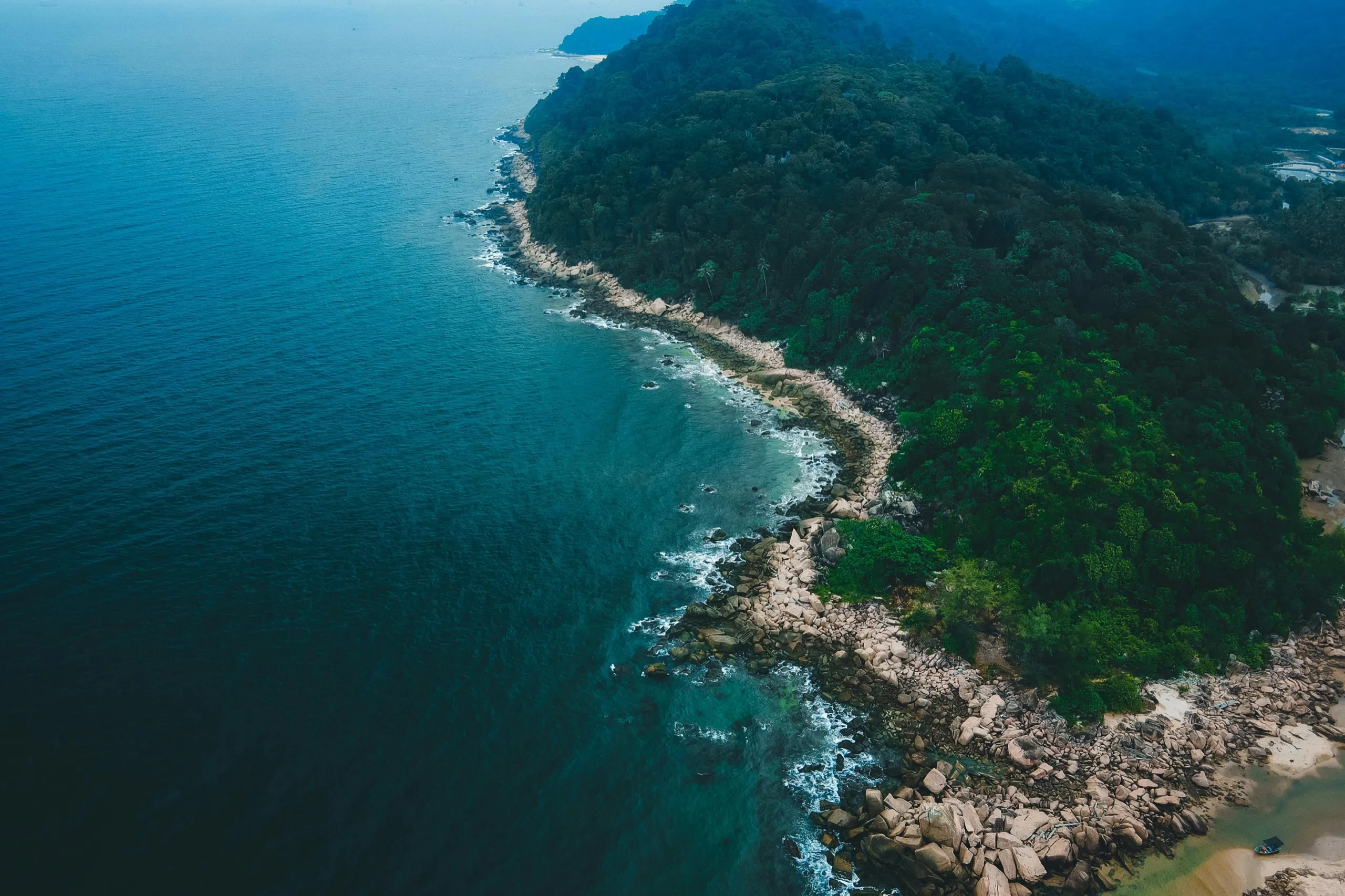 Island in Terengganu, Malaysia. Aerial view of a lush green coastline with rocky shores meeting a calm blue ocean. Dense forest covers the land, creating a tranquil scene.
