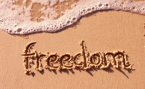 The word “freedom” is written in sand on a beach, with waves approaching. The scene conveys a sense of tranquility and openness.