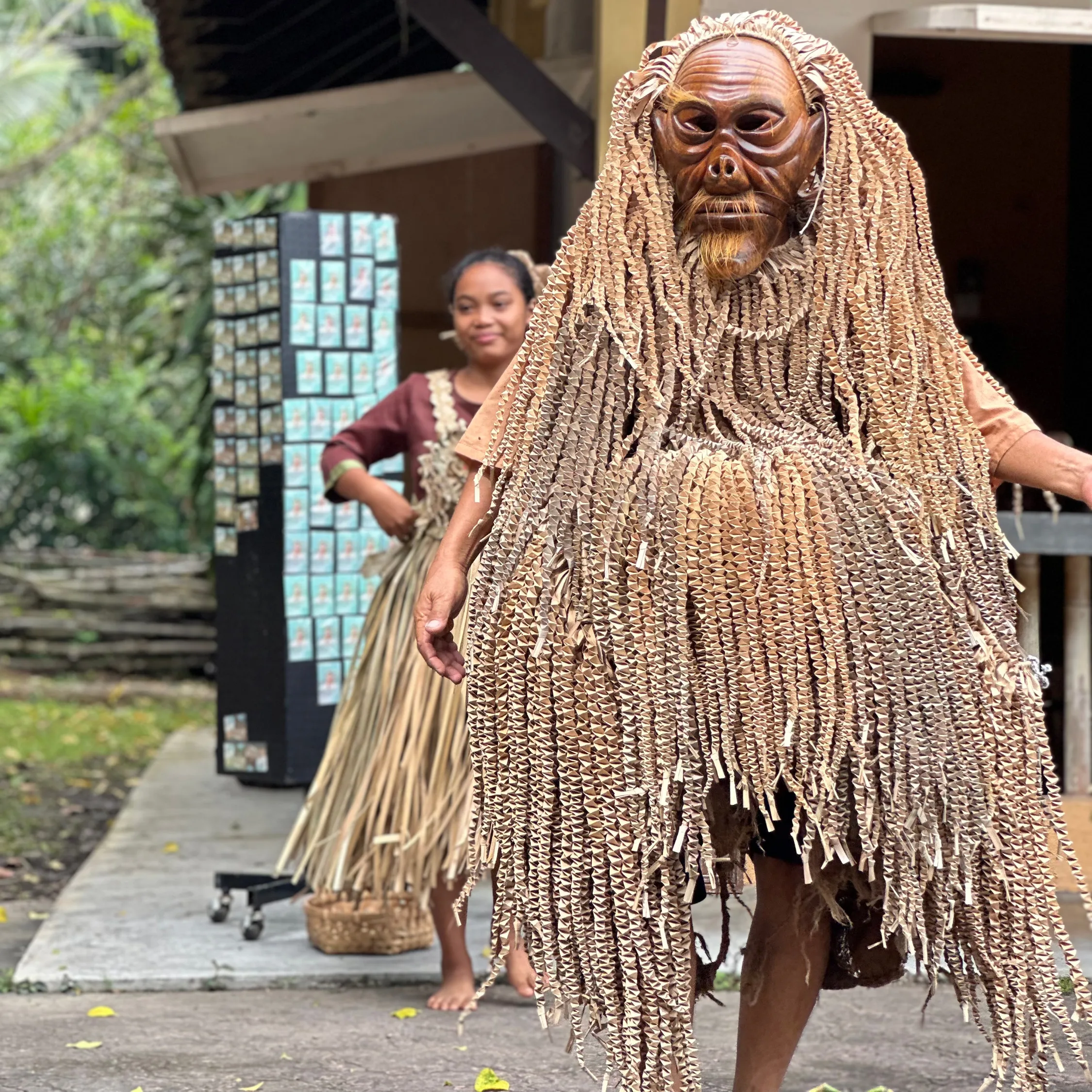 A Mah Meri Tribe person in a detailed wooden mask and braided attire walks forward. Another person in a similar skirt is in the background on a lush path.