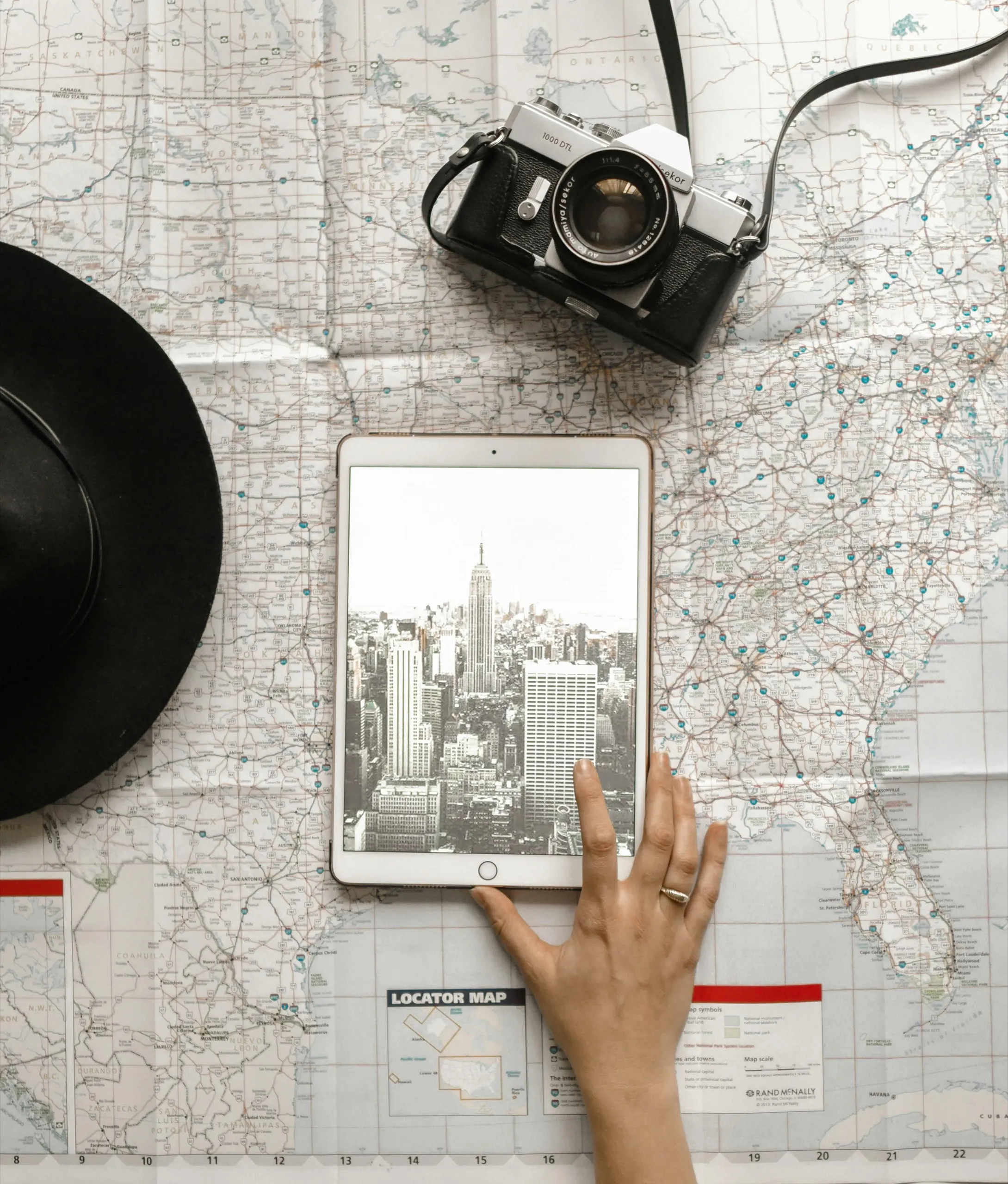 A hand rests on a map with a tablet displaying a city skyline. A vintage camera and a black hat are nearby, creating a sense of travel.