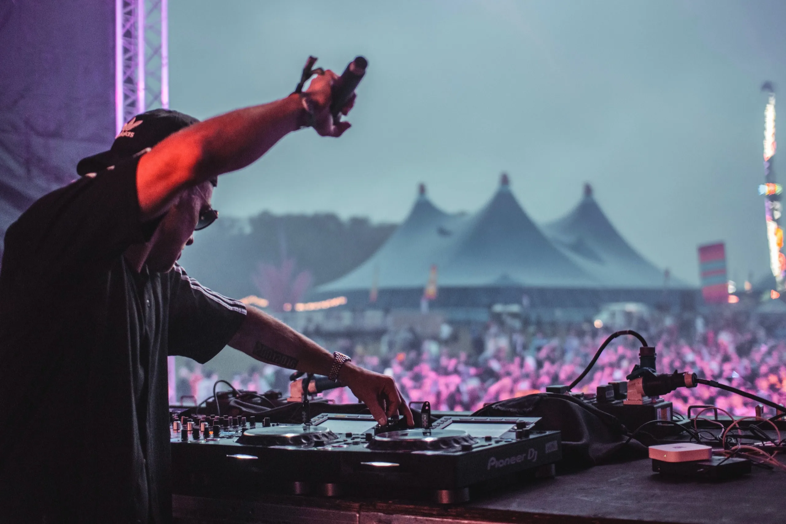 DJ performing on stage with a crowd and festival tents in the background. The music festival is vibrant with purple lighting and an energetic mood.