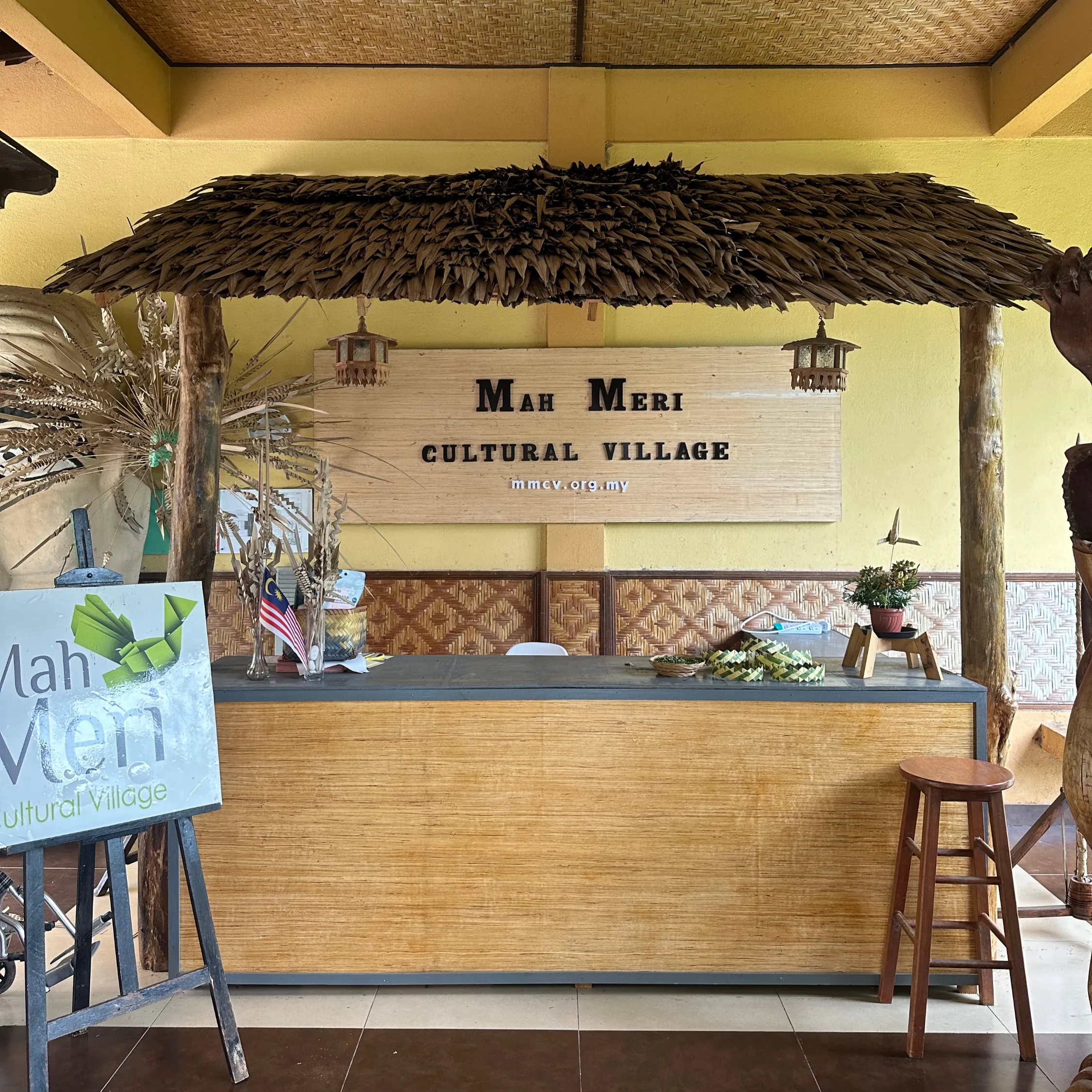 Reception area at Mah Meri Cultural Village with a thatched roof, wooden desk, and decorative plants. Mood is welcoming.