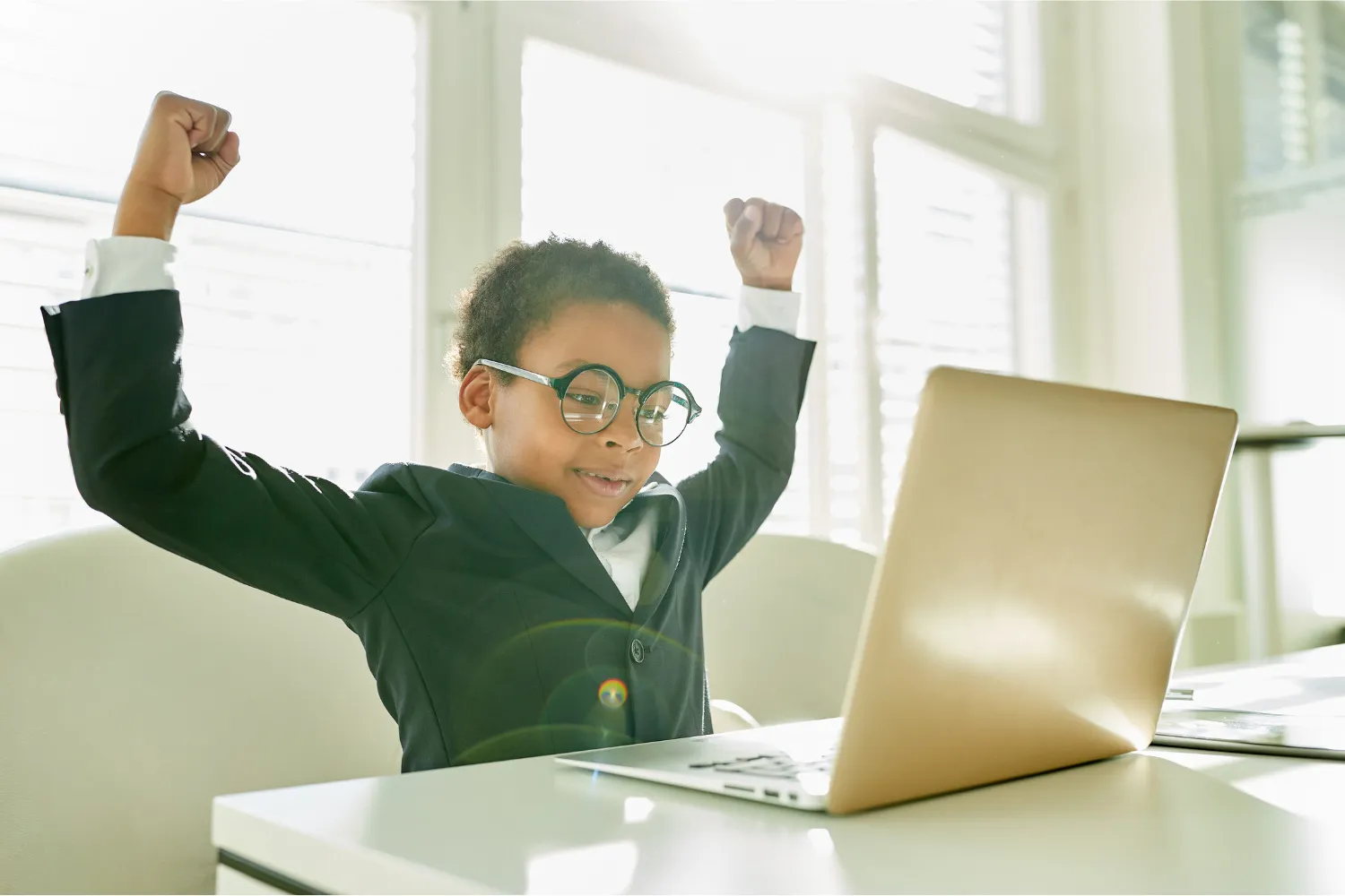 Child in a suit and glasses raises arms in excitement while looking at a laptop. Bright room, sunlight through windows, joyful mood.