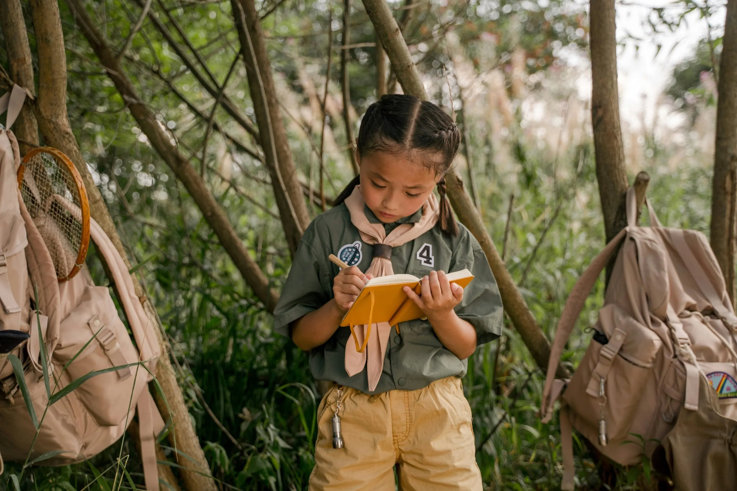 Young girl writing in a yellow notebook in a forest setting, wearing a scout uniform. Backpacks and trees surround her, creating a focused mood.